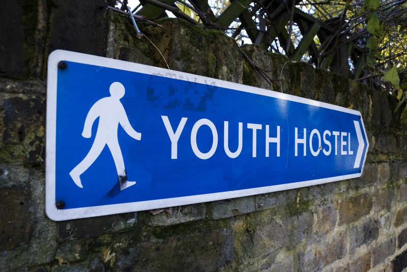 This photo shows a blue sign with white text reading 'youth hostel' and a white icon of a person walking, hanging on a brick wall under green branches, representing the best hostel affiliate programs.