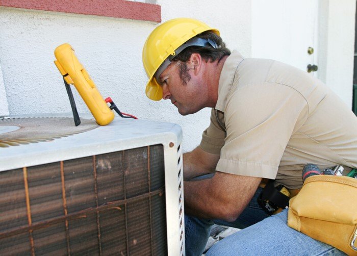 This photo shows a man in a tan shirt, yellow hard hat, and jeans working on an air conditioning unit outside a white stucco building, representing the question: Do HVAC technicians make good money?