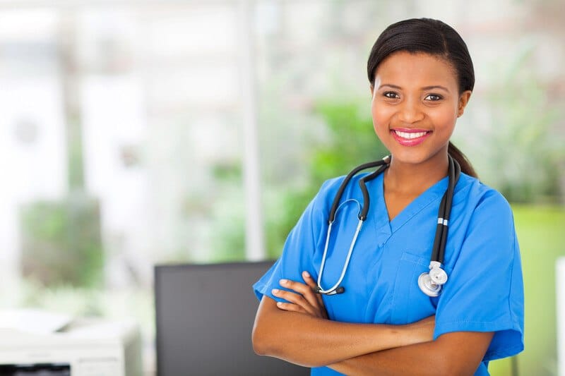 This photo shows a smiling dark-haired woman in blue scrubs standing with her arms folded in front of a window, representing the question, do nurses make good money?