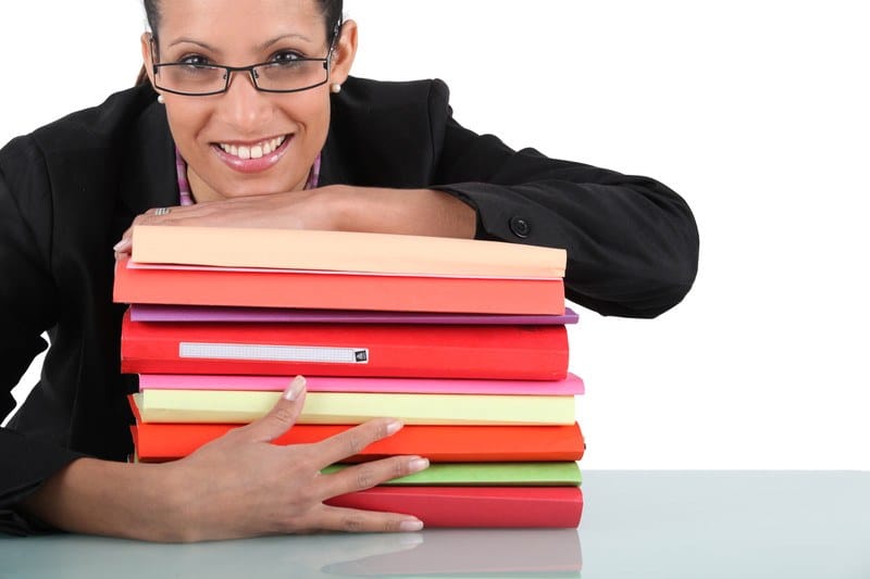 This photo shows a smiling woman in glasses and a dark business jacket leaning across a stack of legal document binders in shades of red and orange, representing the question, do paralegals make good money?