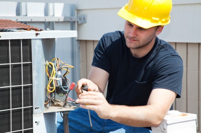 This photo shows a young man in a yellow hard hat, black tee shirt, and jeans working on a the wiring section of an air conditioning unit outside a brown and white building, depicting an HVAC technician at work.