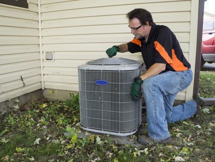 This photo shows a repairman in jeans and an orange and black shirt fixing an air conditioning unit outside a cream-colored home.