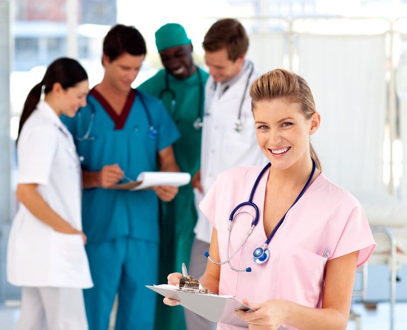 This photo shows a smiling blonde woman in pink scrubs, holding a clipboard, standing in front of a group of doctors and nurses in white, blue, and green clothing, in a hospital setting, representing the idea that nurses have good job satisfaction.