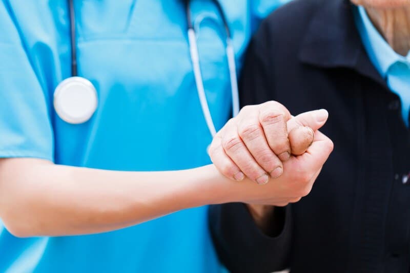 This photo shows a neck-to-waist shot of a woman in a blue nursing uniform holding the hand of an elderly woman in a dark jacket, representing a nurse at work.