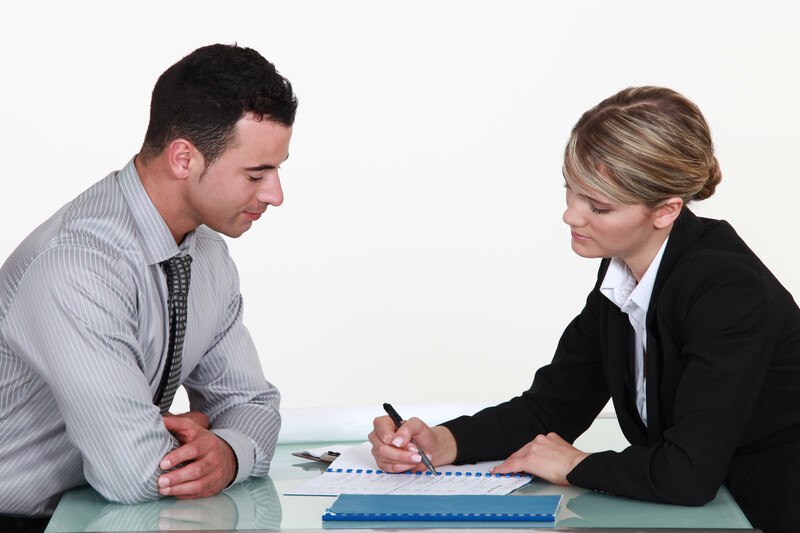 This shows a man and a woman in business clothing leaning over some documents lying between them on the table, while the woman points to something on one of the documents with her pen.