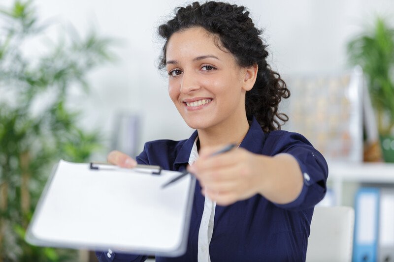 This photo shows a smiling dark-haired woman in white and navy blue business clothing holding out a clipboard and pen.