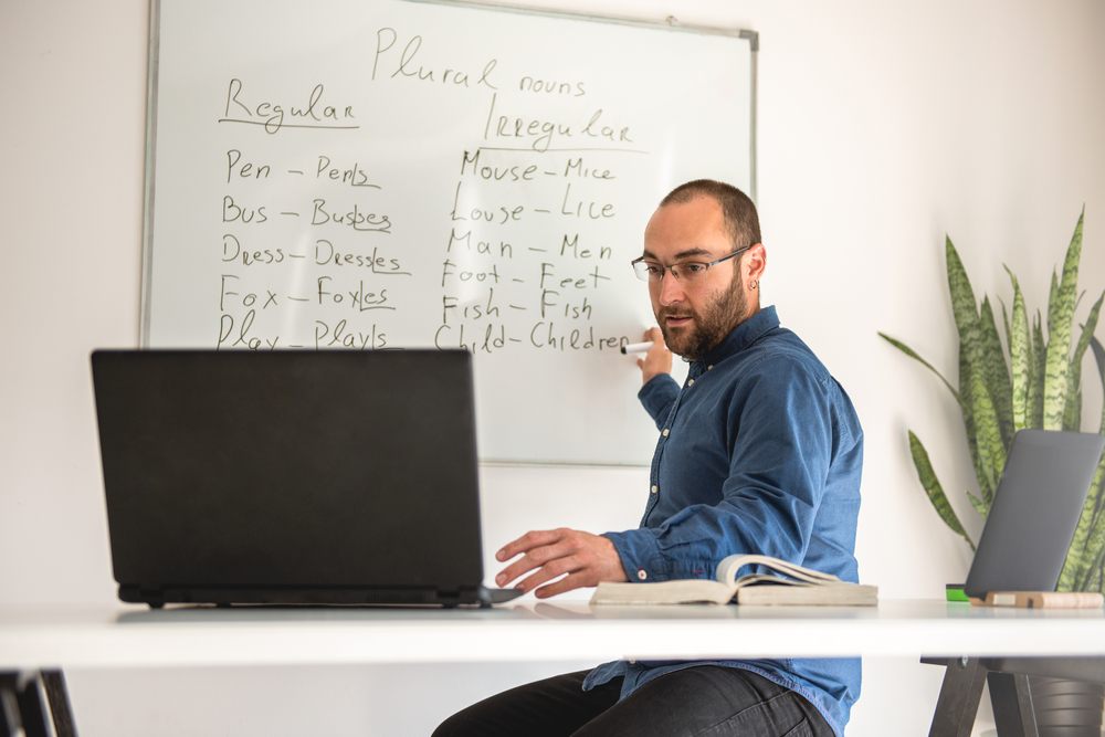 adult man with beard teaching English on his laptop with white board