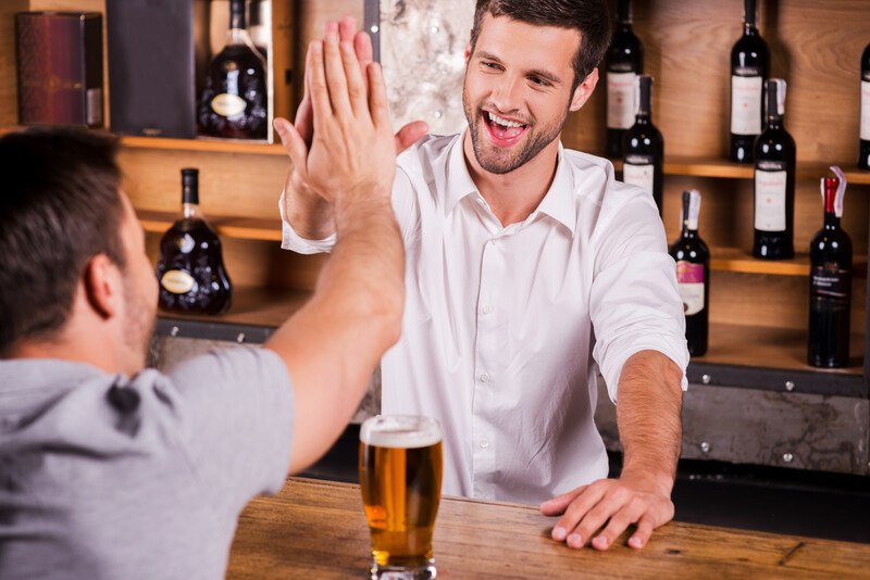 This photo shows the rear perspective of a man in a gray shirt sitting a bar, giving a high five to a smiling bartender in a white shirt, representing a bartender at work.