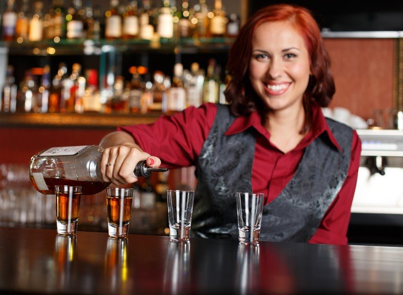 This photo shows a pretty readheaded bartender in a red blouse and gray vest pouring shots on a dark wooden bar.