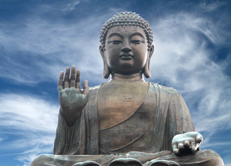 This photo of the Tian Tan Buddha statue in Hong Kong has a bronzed look and sits before a dramatic blue sky with white feathery clouds, representing the best Buddhism Affiliate Programs.