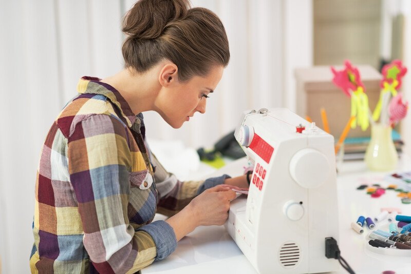 This photo shows a brunette woman in a bun and a plaid shirt using a sewing machine in what appears to be a craft room, representing the best handmade craft affiliate programs.