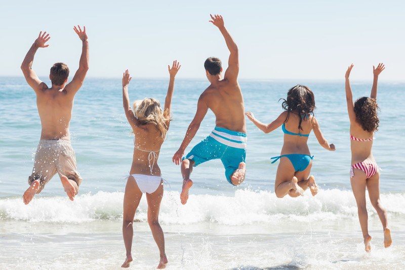 This photo shows a back view photo of a group of friends in varying colors of swimsuits playing in the water at a beach, representing the best swimwear affiliate programs.