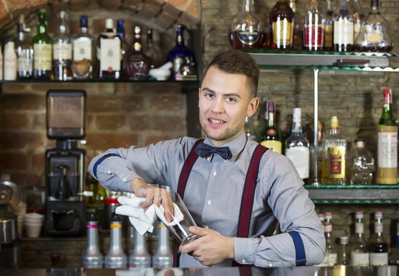 This photo shows a handsome young bartender in a gray shirt, red suspenders, and blue bowtie wiping a glass behind a bar, representing the question, do bartenders make good money?