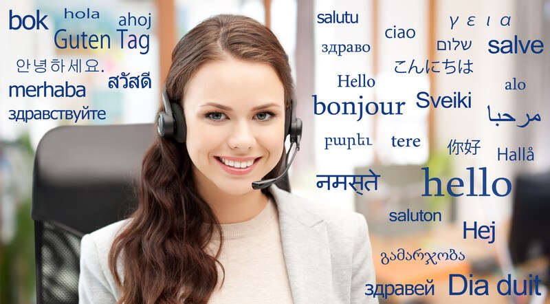 This photo shows a smiling brunette woman with a headset and light-colored clothing sitting in an office chair surrounded by words in blue text meaning 'hello' in several languages, representing the question, do translators make good money?