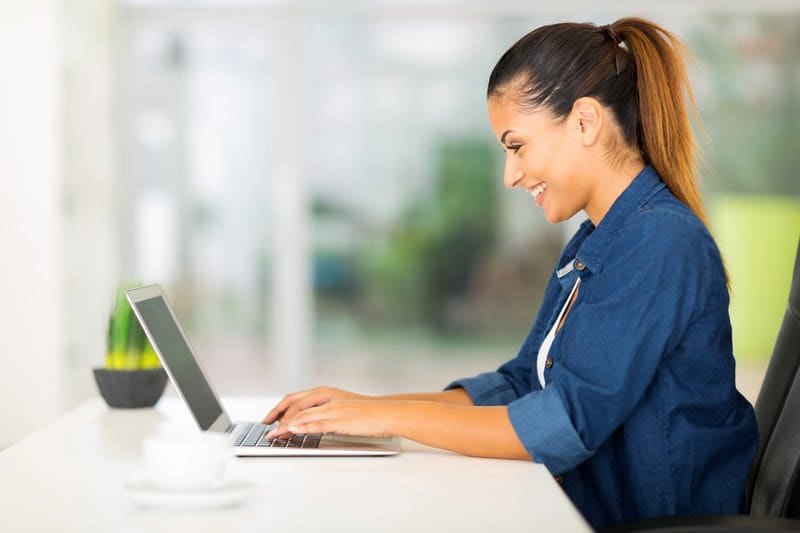 woman in pony tail and blue casual clothes working on her laptop at home