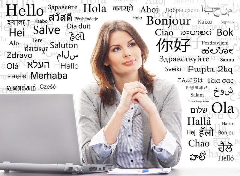 This photo shows a brunette woman in a gray sweater and blue and white striped shirt leaning forward on her elbows on a table next to a laptop and a paper with a pen, surrounded by words in black text in several languages.