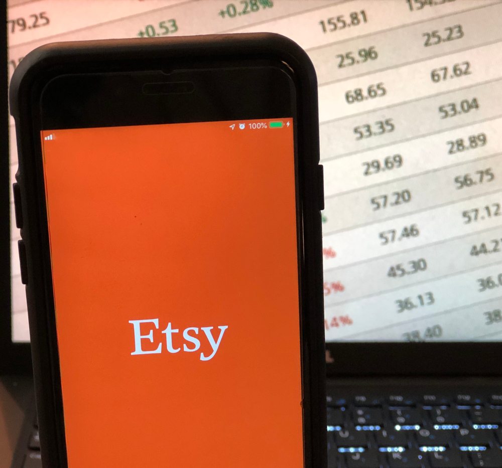 close photo of etsy app on smartphone with sale prices on laptop in background