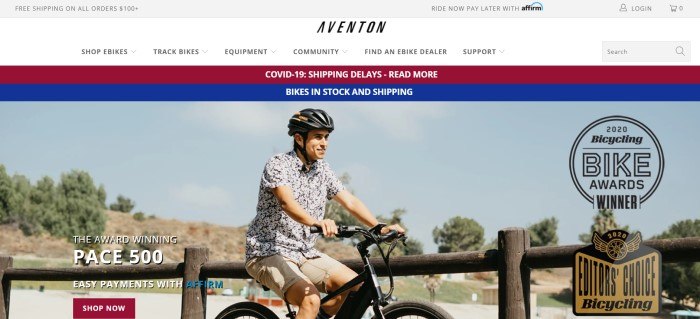 This screenshot of the home page for Aventon has a gray header, a white navigation bar, and a red and blue announcement bar above a photo of a smiling man in a black helmet, a black and white print shirt, and khaki shorts riding a black bike in what appears to be a park, along with white text and a red call to action button.