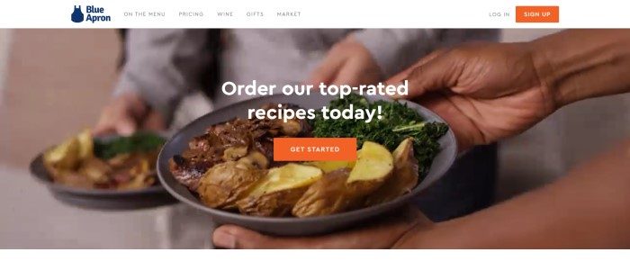 This screenshot of the home page for Blue Apron has a white header with black text, a blue logo, and an orange log-in button on the navigation bar, above a photo of two people carrying plates of grilled meat, potatoes, and spinach behind white text and an orange call to action button.