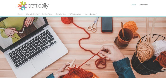 This screenshot of the home page for Craft Daily has a white header with a multicolored logo above an overhead photo showing a laptop with a knitting demonstration on the screen, two balls of orange yarn, a pair of scissors, a mobile phone, and an orange coffee cup resting on a wooden table, as well as a pair of hands holding knitting needles.