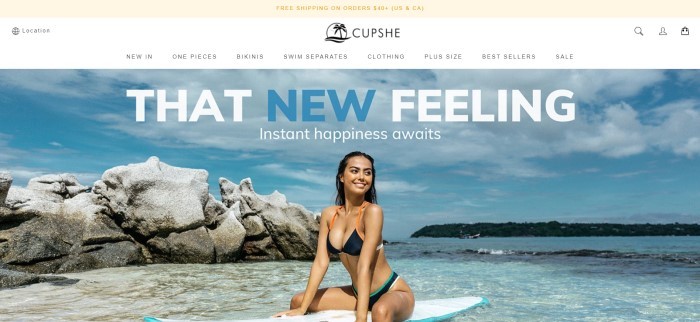 This screenshot of the home page for Cupshe has a beige header, a white navigation bar, and a large photo of a smiling dark haired woman in a back bikini sitting on a surf board in the ocean, behind white and blue text.