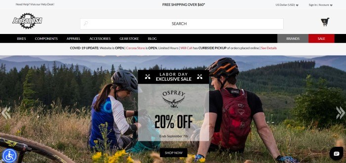 This screenshot of the home page for Jenson USA has a gray header and search bar, a black navigation bar with white text, a white announcement bar, and a large photo showing the rear view of a young man and young women in bicycle clothing and helmets sitting in a meadow near their bikes, opening their backpacks as they look over an evergreen forest with mountains in the background, along with a gray and black sales announcement and a black call to action button.