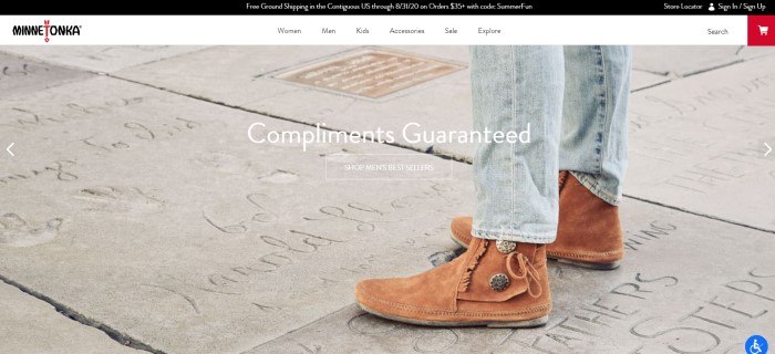 This screenshot of the home page for Minnetonka has a black header, a white navigation bar, and a photo showing a man's legs in jeans and brown suede Minnetonka boots standing on concrete that had been written in, along with white text and call to action button.