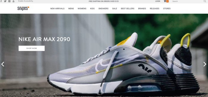 This screenshot of the home page for Snipes has a gray header, a white navigation bar with black text, and a photo of a silver, yellow, white, and black sneaker lying on a sidewalk near a white fence, along with white text and a white call to action button on the left side of the page.