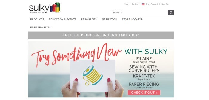 This screenshot of the home page for Sulky has a white background and black text through most of the page, a gray announcement bar below the navigation section, and a photo showing a woman's hands holding a card with a graphic of thread behind red, green, and black text, as well as a red call to action button.