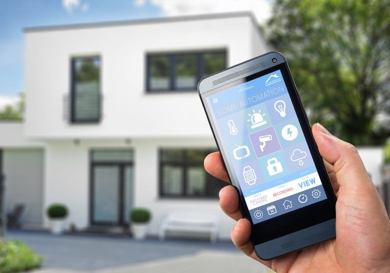 This photo shows a white home in the background with a man's hand holding a mobile device with home connection icons on the screen in the foreground, representing the best smart home affiliate programs.