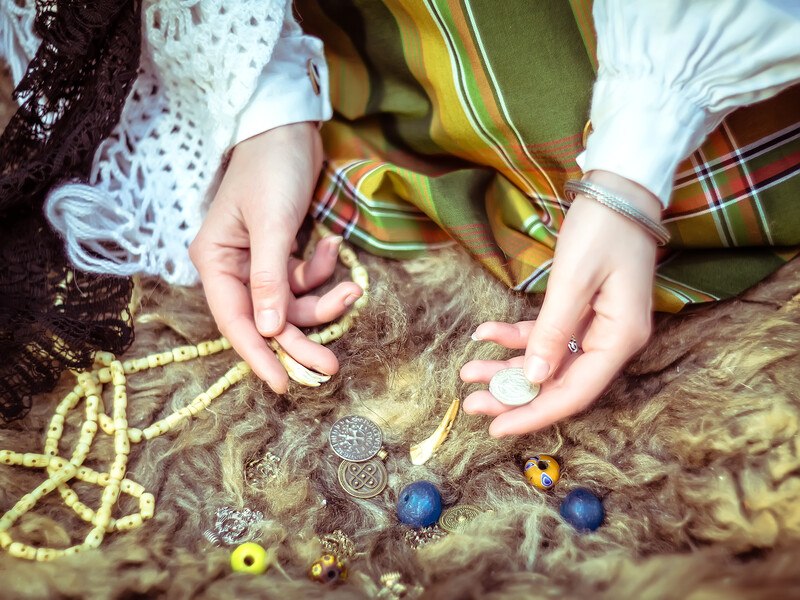 This photo shows the hands of a young woman sorting through various beads, coins, and runes as if preparing to cast a spell, representing the best witchcraft affiliate programs.