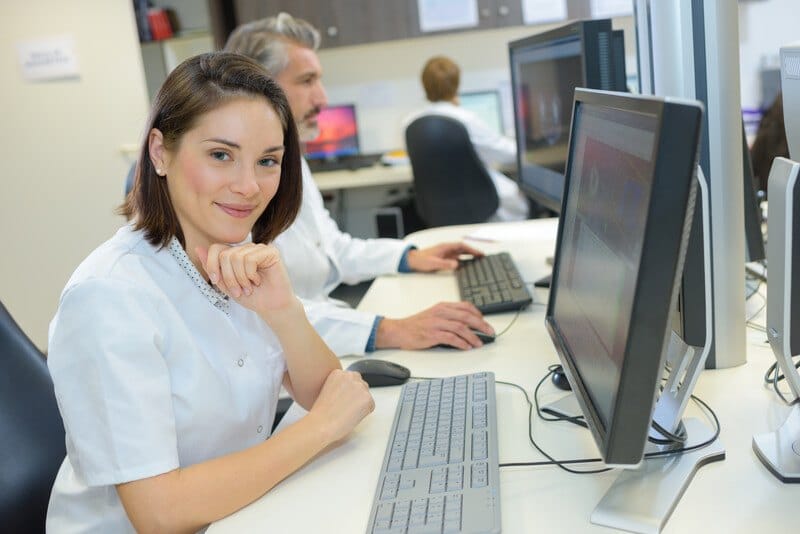 This photo shows a smiling brunette woman in a white shirt leaning on her desk near a keyboard and monitor in an office setting with other workers, representing the question, do medical coders make good money?