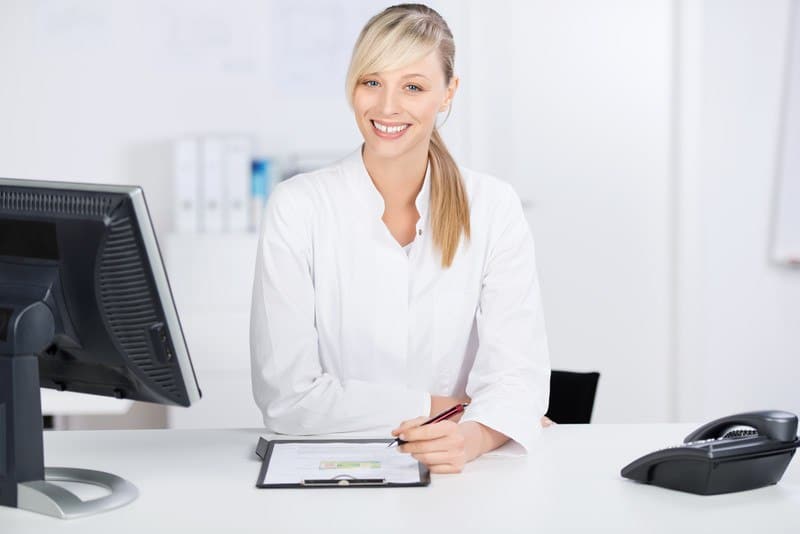 This photo shows a smiling blonde woman in a white shirt holding a pen near a desk with a phone, a monitor, and a clipboard with medical information on it.