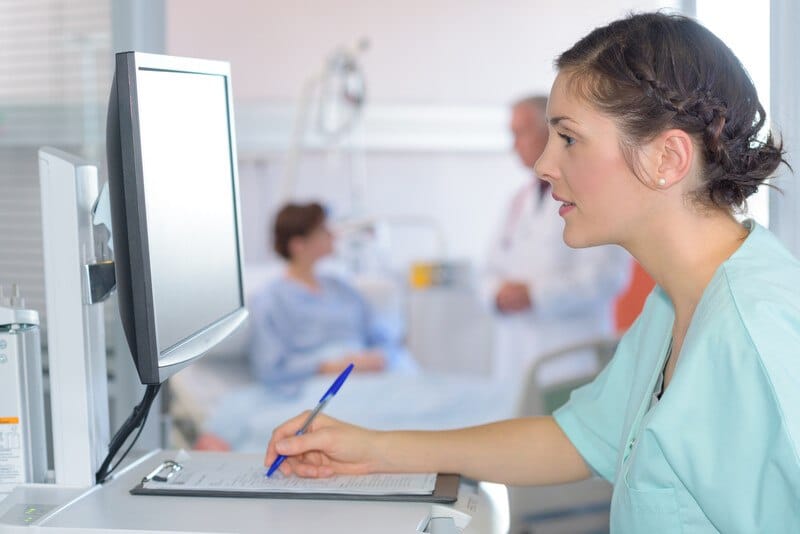 This photo shows a brunette woman in scrubs focusing on a computer screen as she holds a pen over a clipboard with medical information on it.