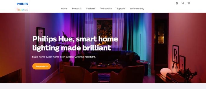 This screenshot of the home page for Philips-Hue has a white navigation bar with black text above a large photo of an indoor living room with lighting set to red, blue, green, and purple, along with white text and an orange call to action button.