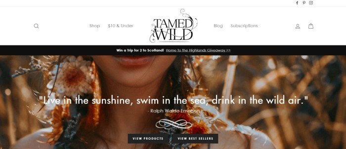 This screenshot of the home page for Tamed Wild shows a black header and a closeup of the chin and shoulders of a woman with long flower-twined hair, behind white text and two black call to action buttons.