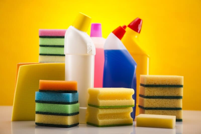 This photo shows several multicolored cleaning bottles and sponges on a glossy white surface in front of a bright yellow background, representing the best cleaning products affiliate programs.