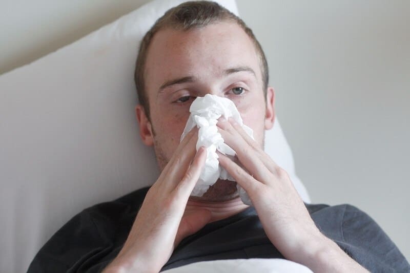 This photo shows a bleary-eyed man in a gray shirt lying on a white pillow while he holds white tissues over his nose and mouth, representing the best cold & flu affiliate programs.