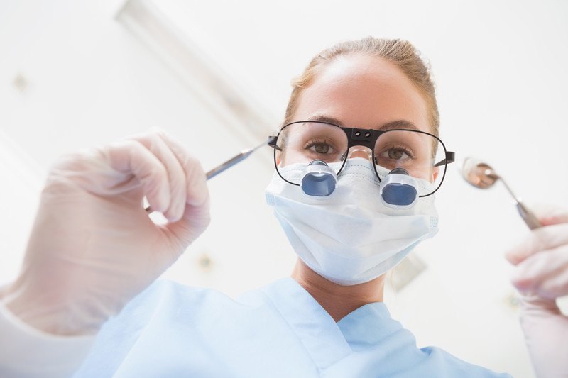 This photo shows a view of a woman in glasses, a face mask, light blue scrubs, and gloves, leaning forward with dental instruments in her hands.