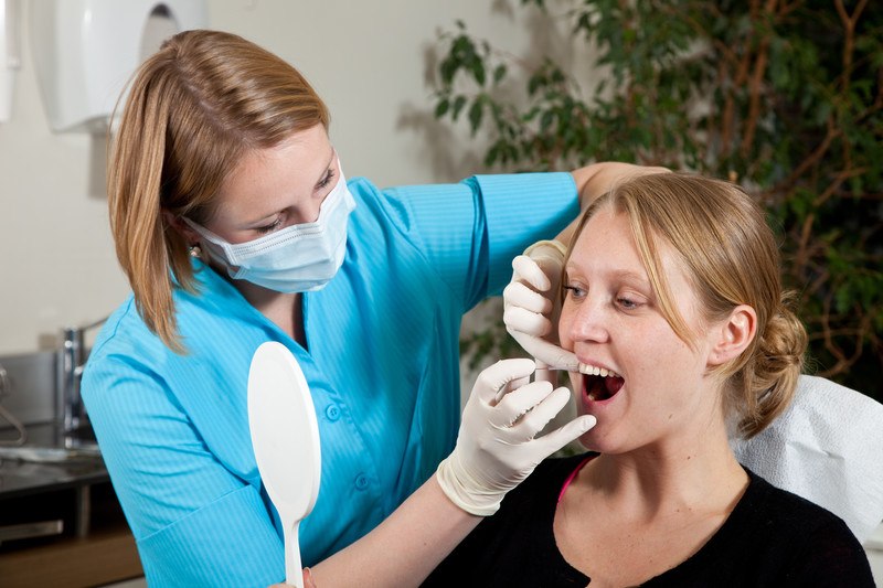 This photo shows a dental hygienist in a turquoise shirt teaching a blonde woman in a dental chair how to floss properly.