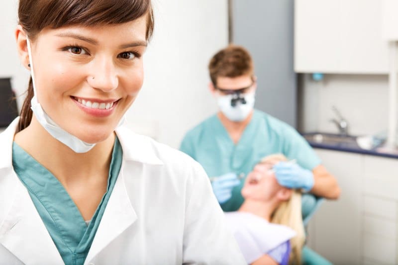 This photo shows a smiling brunette woman in aqua scrubs and a white lab coat standing in a dentist office in front of a blonde woman lying in a dental chair with her mouth open, receiving an oral exam from a dentist in aqua scrubs, representing the question, do dental hygienists make good money?