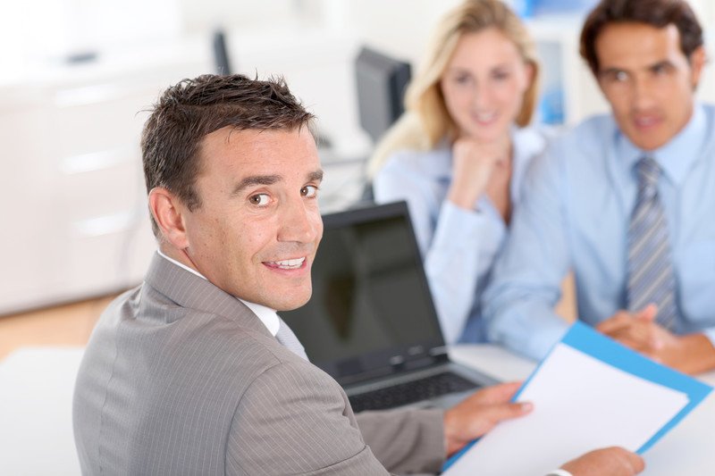 This photo shows a smiling dark haired man in a gray suit near an open laptop holding some paperwork toward a blonde woman and a dark-haired man sitting across a white table from him, representing the question, do loan officers make good money?