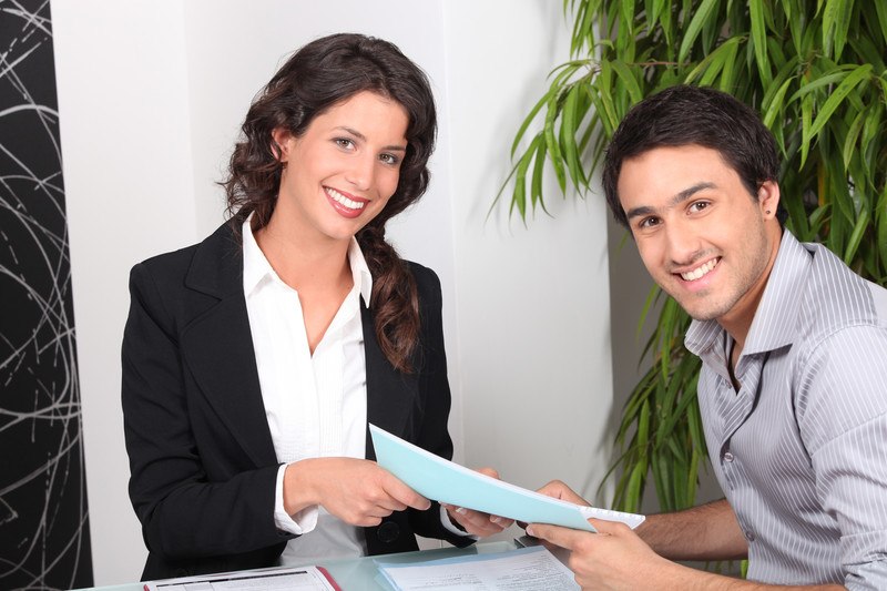 This photo shows a smiling dark haired woman in a black suit and white blouse handing some paperwork to a smiling dark haired man in a gray and white striped shirt.d