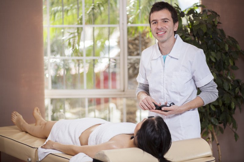 This photo shows a smiling brunette massage therapist preparing to work on a woman in a white towel lying on a table near a window.