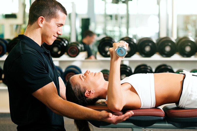 This photo shows a male personal trainer in black clothing helping a female client in white workout clothing as she lifts weights.