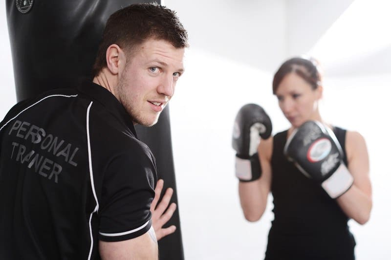 This photo shows a male personal trainer in black helping a female client in black work out with a punching bag and gloves.