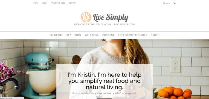 This screenshot of the home page for Live Simply has a white header and navigation bar with black text and a black and pink logo above a large photo showing the neck and torso of a woman with long blond hair and a tan sweater standing in a white-tiled kitchen with purple flowers, a bowl of oranges, and a blue stand mixer, behind a white text box with black text.