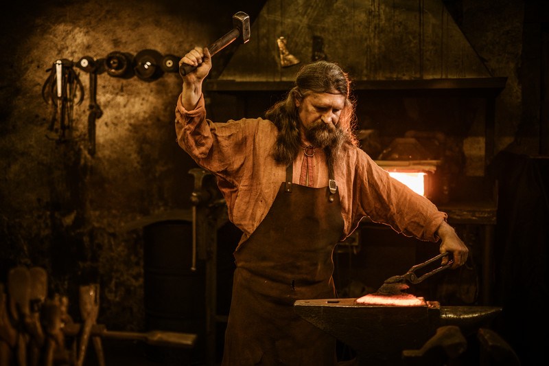 This photo shows a blacksmith in a forge swinging a hammer over his head as he aims toward a piece of glowing metal he is holding on an anvil with a pair of tongs, representing the best blacksmith affiliate programs.