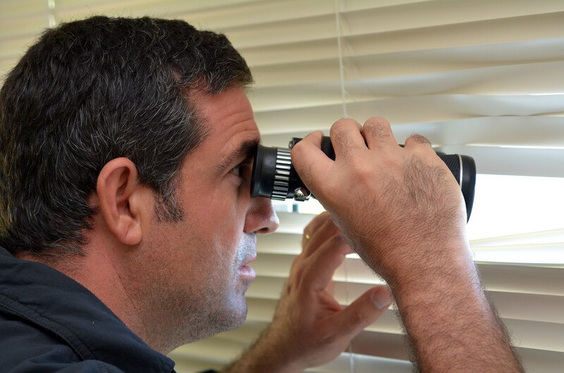 This photo shows a dark haired man in a dark shirt looking with binoculars through the blinds of a window, representing a bounty hunter at work surveilling a fugitive.