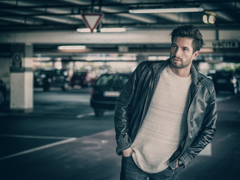 This photo shows a bearded young man in jeans, a white top, and a black jacket in an underground parking lot, representing a bounty hunter looking for a fugitive.